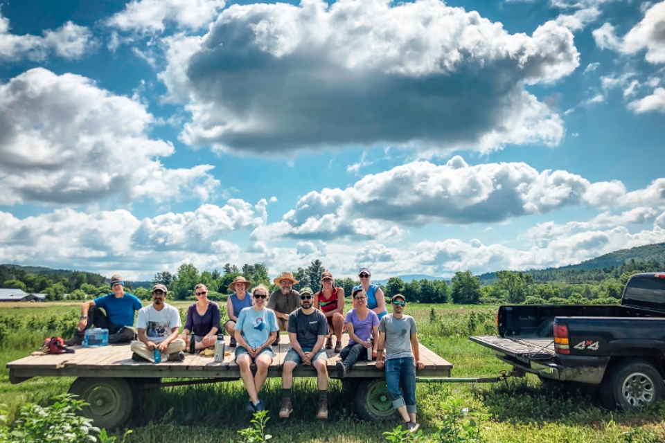 A group of City Market Members sitting on a flat trailer hitched to a green pickup truck. The truck is parked in a lush green field. The top half of the photo is rich blue sky dotted with fluffy clouds that are lit from above, with white tops and dark gray bottoms.