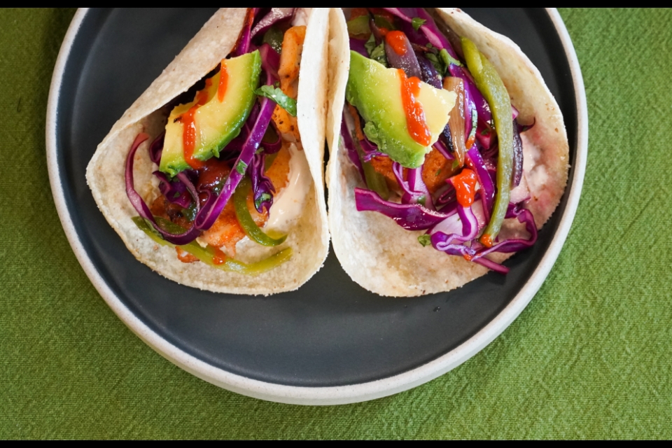 Two fish tacos on a slate gray plate. The tacos are topped with green avocado, purple cabbage slaw and orange hot sauce.