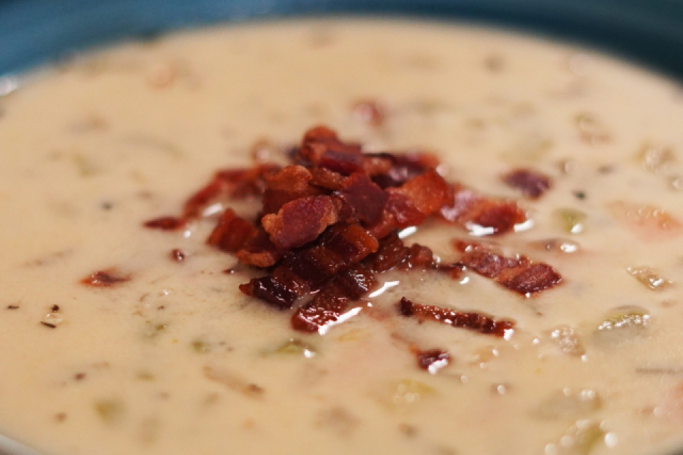 A close up of the soup in a teal bowl. The soup is white, with bits of celery, carrot, and onion floating within. Topping the center of the soup is a pile of crumbled bacon. 