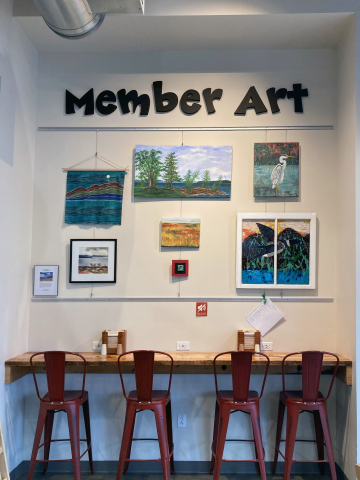 A counter with four red high-top chairs facing away from the viewer. The counter is in front of a wall with large black lettering at top that reads "Member Art." Below the lettering, several paintings of different sizes and many colors hang in three columns.