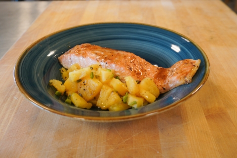 Baked salmon with glaze on it next to yellow pineapples and cut up green jalapenos. The salmon is plated on a plate with dark and light blue rings and a brown rim, placed on a cutting board.  