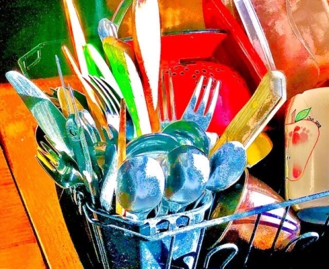 Photo of art by Christine Henninger. It features a close-up of highly saturated silverware in a dish drying rack, with a backdrop of other dishes including a red collander and a mug with a minimalist drawing of an apple cross-section.