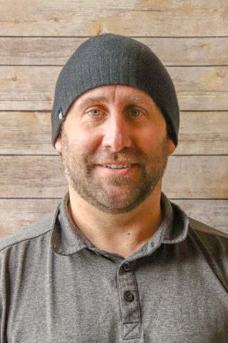 A photo of a person from the shoulders up in front of a wood-panel backdrop. The person is wearing a gray polo shirt and a darker gray knit beanie. He has a slight smile.