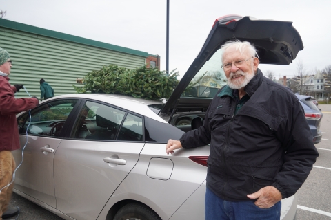 A man stands behind a silver sedan. He has white hair and a white beard, and is wearing wire framed glasses. He is wearing a black coat and has one hand tucked into the pocket of his jeans.