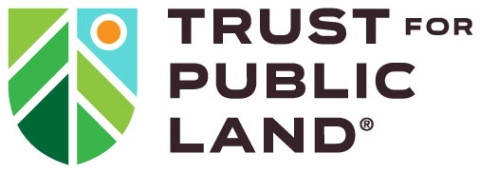 The logo for the Trust for Public Land consists of a shield split in half. The left side has green diagonal bars that evoke forest. The right side has a blue triangle at top with a yellow circle in the middle, then green bars below, evoking a sky over a forest. To the right of the shield, "Trust for Public Land" is printed in all black capital letters.