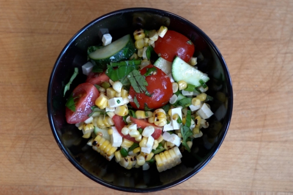 A top-down view of a black bowl with a completed grilled corn and zucchini salad with vibrant red cherry tomatoes, white ricotta salata cheese, and green chopped basil against a wooden cutting board.