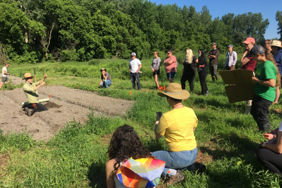 12 people are arranged in a semicircle at the right of the photo. Some are sitting and some are standing on a grassy field. To their left, a person is sitting in a field of dirt. They are wearing a green shirt and brown hat, and they are holding a sapling.