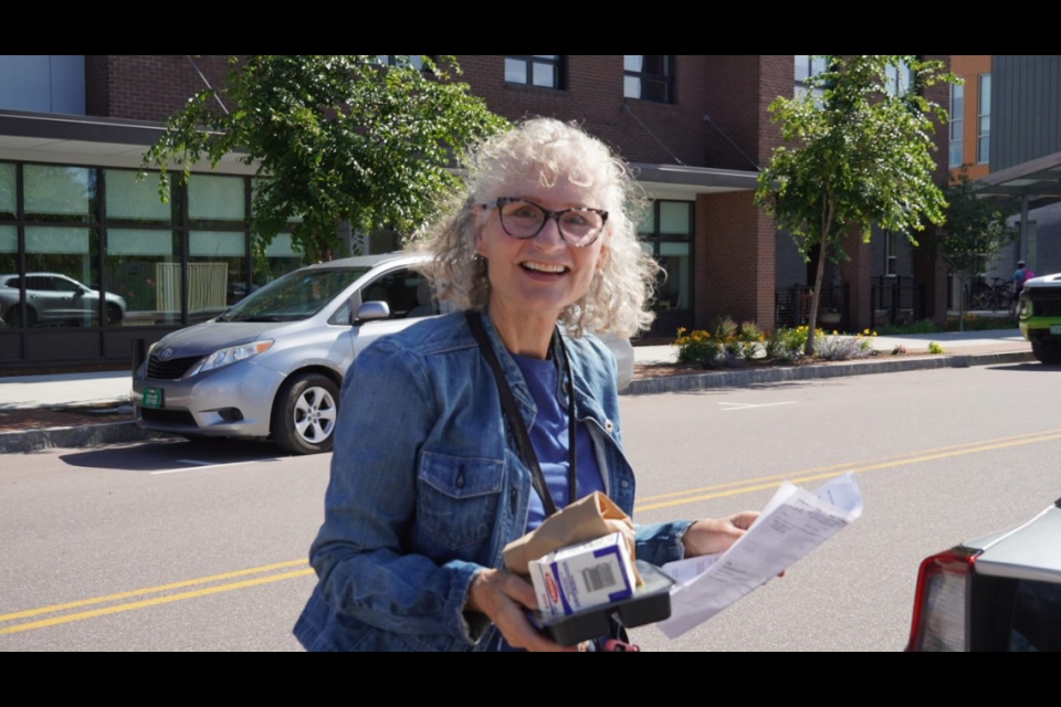 A woman with curly white hair and black rimmed glasses stands in front of a street. She is wearing a blue denim jacket, holding a few sheets of paper, and smiling widely.