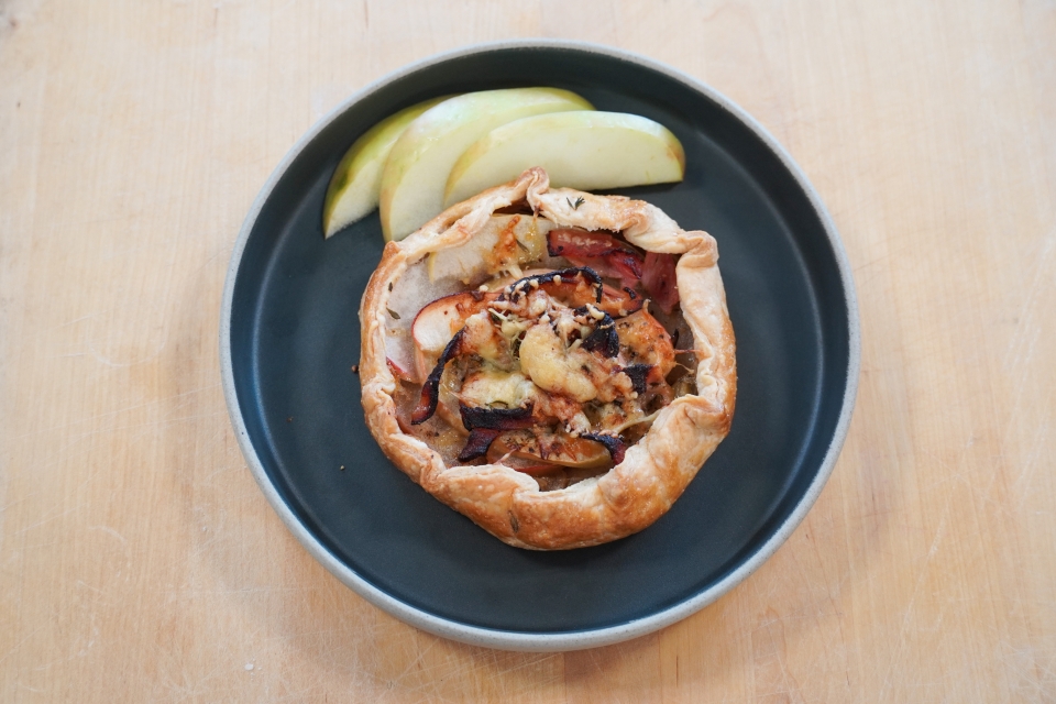 Apple galette with crisped ham, baked apples, and melted cheese in a pie crust. Three sliced honeycrisp apple pieces behind it on a black plate that sits on a lightwood cutting board.