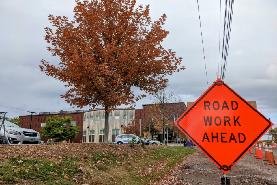 A bright orange diamond-shaped sign reads "Road Work Ahead." Behind it, a tree with a full, dusty orange crown stands in front of the City Market store in the South End. The sky is grey-blue.