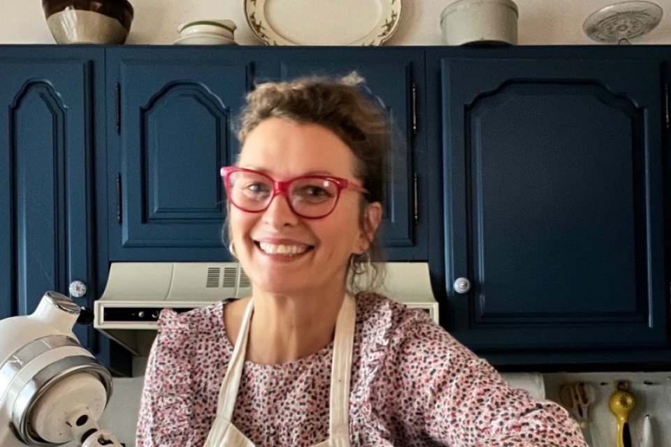 A woman wearing red glasses stands near a mixer and baking ingredients in a kitchen with blue cabinets