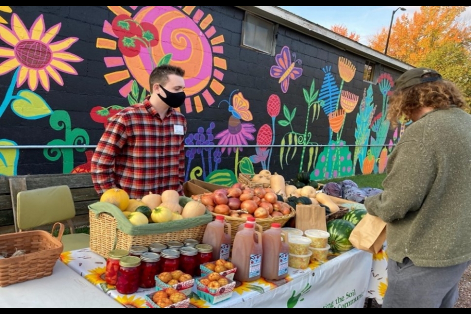 A person stands in front of a mural that is painted on the side of a building. The mural has a black background and features brightly-colored flowers and insects. The person is wearing a red plaid shirt and a black mask, and has a table in front of them that is piled high with various squash, apples, cider, and greens.