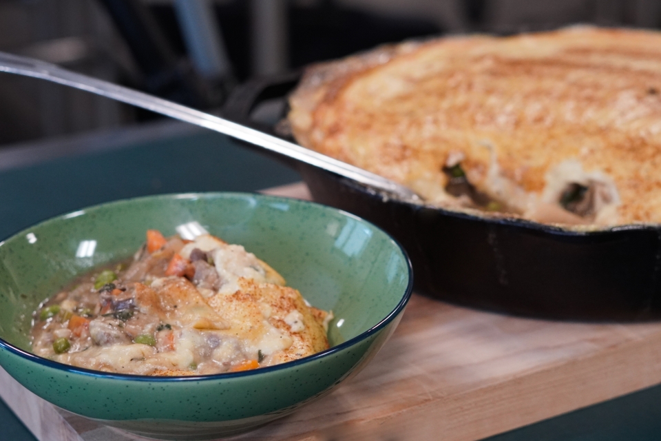 Green bowl with navy lining with carrots, peas, stock, and polenta with cheese inside of it. The bowl sits on a cutting board with rest of cottage pie in background with metal serving spoon out of black cast-iron pan.