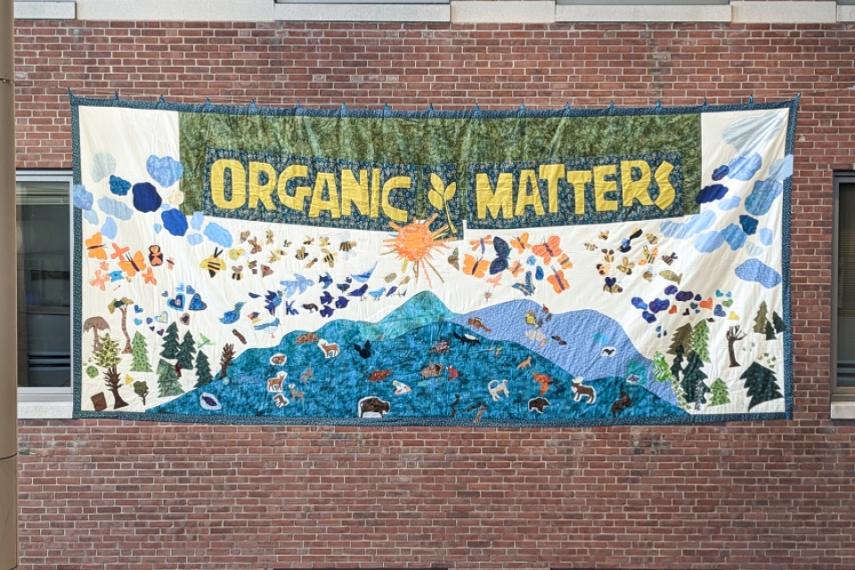 A brick wall with a quilted banner hung on it covering some windows. The banner reads Organic Matters in golden-rod colored letters on a green background. The rest of the banner is white with a blue border and teal and pale blue mountains with an orange sun. 