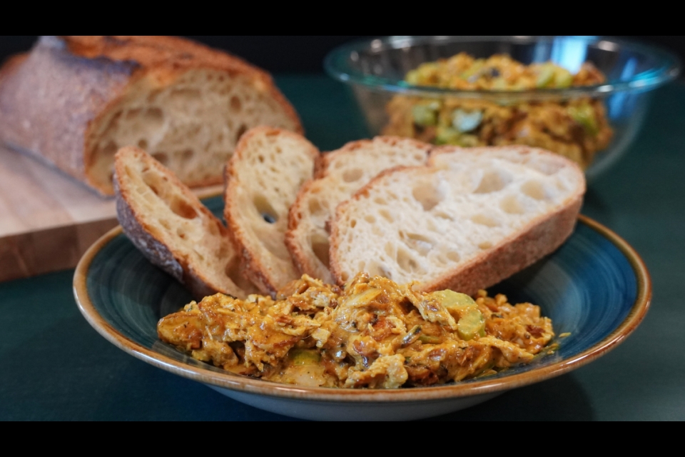Our curry chicken salad is on a plate with four slices of hearty bread piled behind it. Behind that plate to the right is a bowl of chicken salad, to the left a cutting board with the rest of the loaf. 