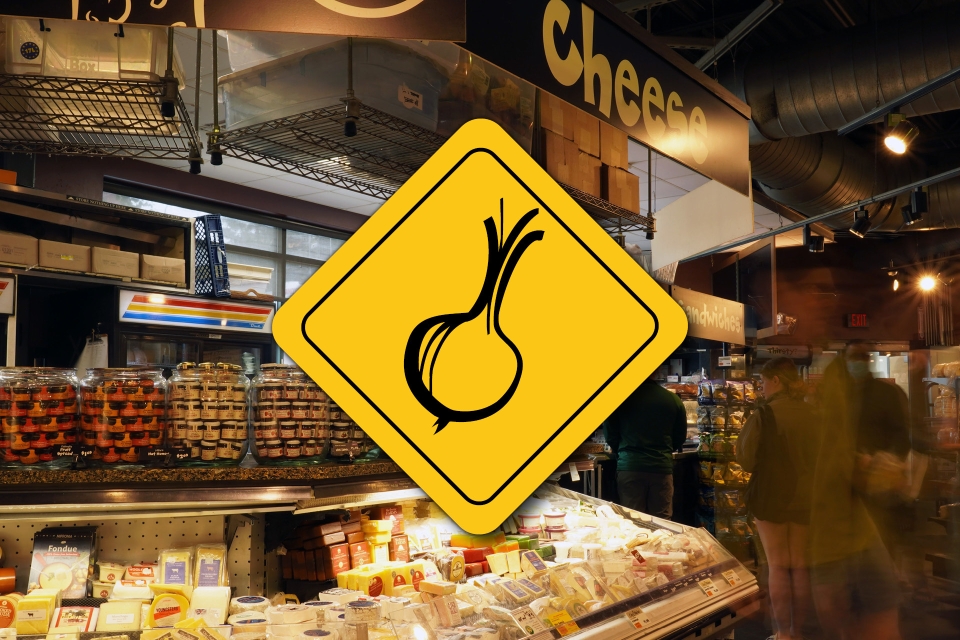 A photo of the cheese department at the Downtown City Market location. There is a yellow diamond with a drawing of an onion on it superimposed over the center of the photo.