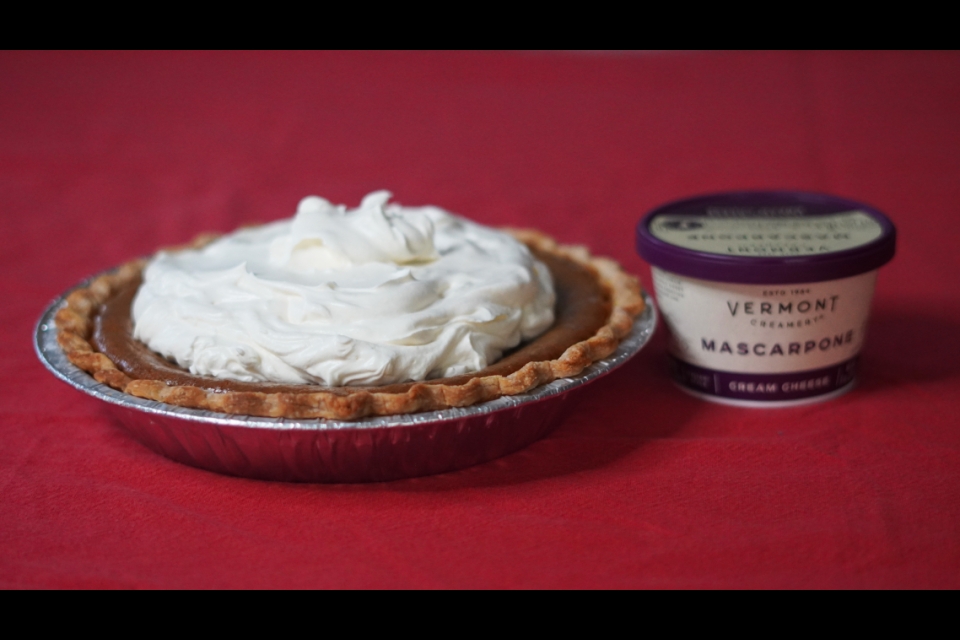 A pumpkin pie with white whipped topping on a red background. Beside the pie there is a container of Vermont Creamery Mascarpone.