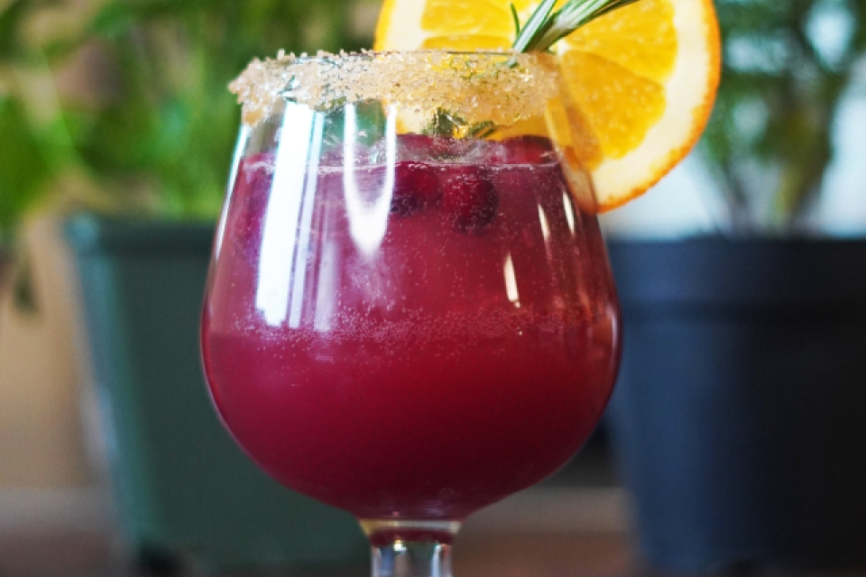 The sunset in a little cocktail glass, there is an orange slice on the rim and a sprig of rosemary sticking out of the glass.