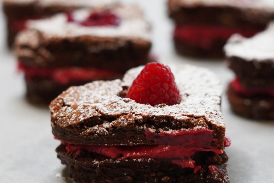 A close up of a heart shaped brownie, with a layer of red cream in the middle, and topped with a raspberry. behind it, and out of focus, are more brownies!