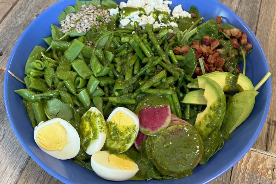 A salad in a blue bowl. Greens are topped with sunflower seeds, watermelon radish, boiled eggs, pancetta, asparagus, goat cheese, avocado, and snap peas