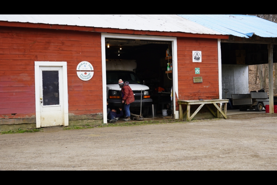 A red barn with a Pomykala Farm sign on the side. There's an open garage door, and through it, a white van is visible. A person in a red coat is working on the front of the van.