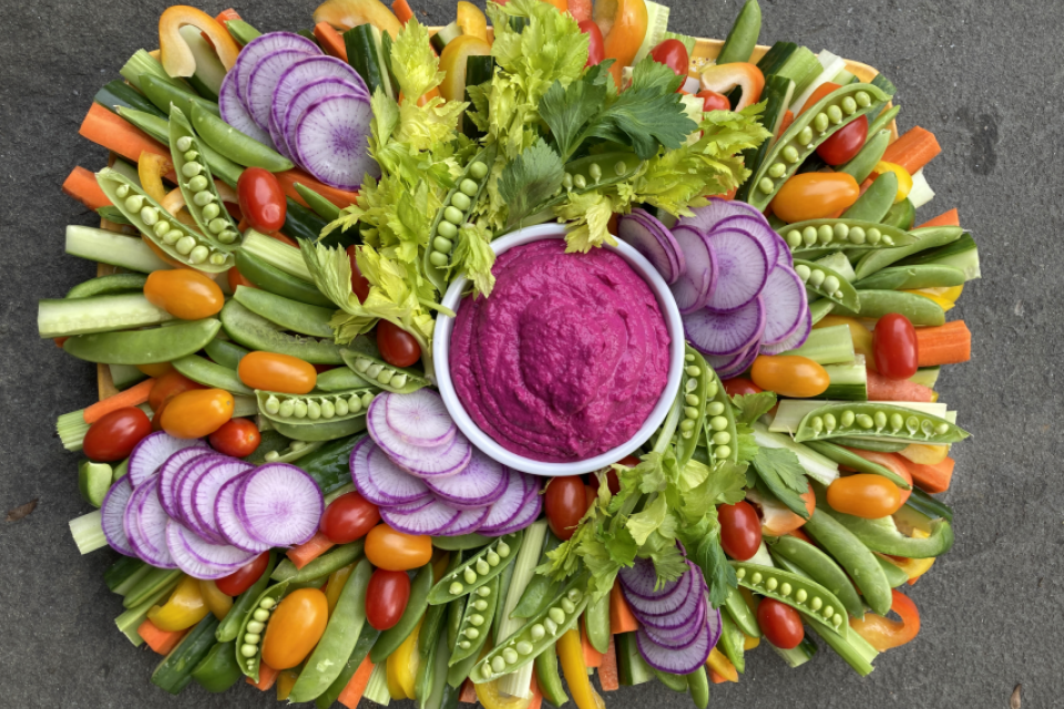 A bountiful collection of sliced vegetables, including snap peas, tomatoes, carrots, daikon, yellow bell peppers, parsley, and celery are arranged around a bowl of bright magenta beet hummus.