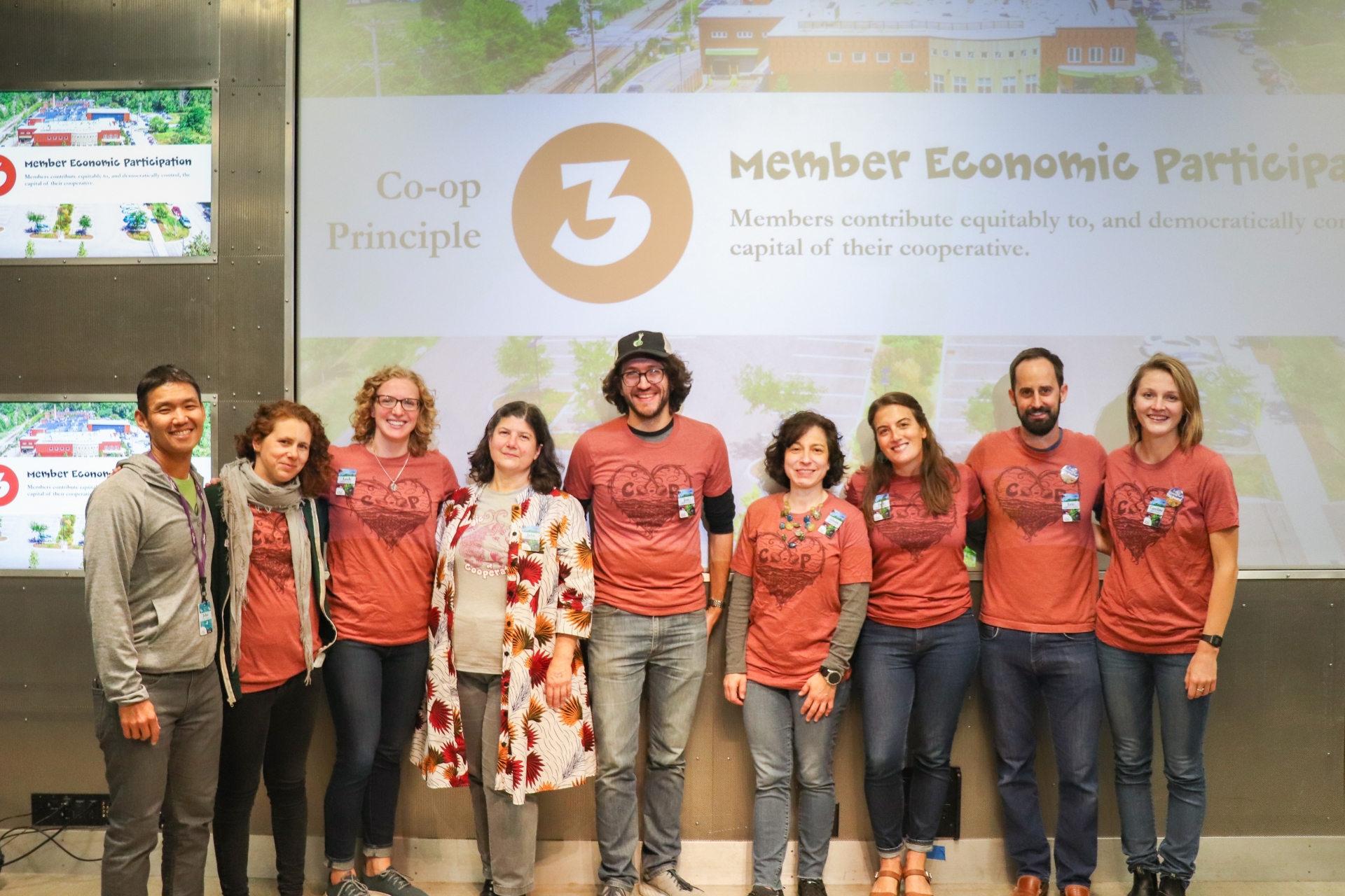 A row of nine people posing in front of a large projector screen. The graphic on the screen is faded and reads "Co-op Principle 3 Member Economic Participation." The number 3 is printed inside a rust red circle. The people
