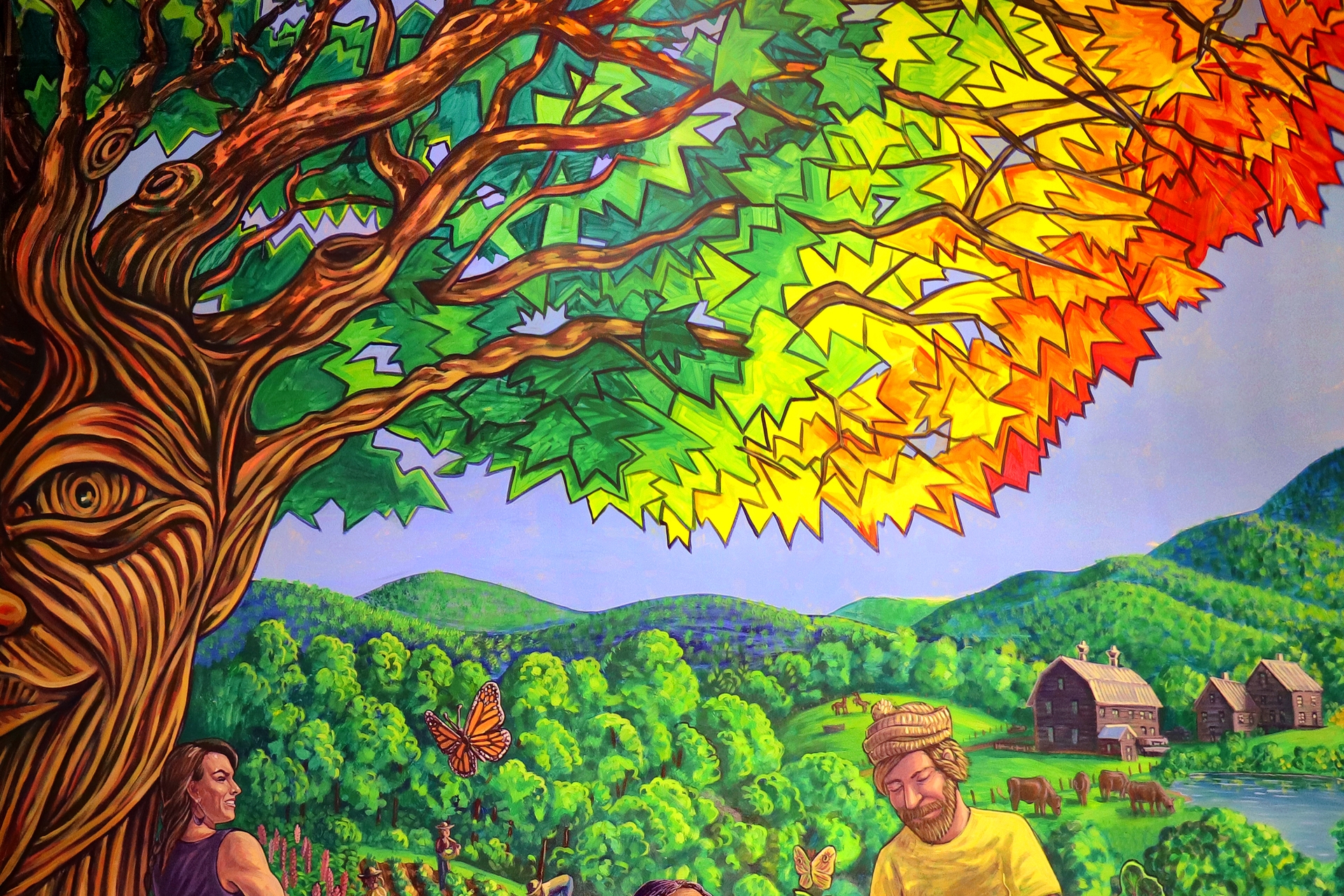 A painting with a tree in the foreground. The tree trunk forms a face. The leaves of the tree nearest the trunk are dark green, but become light green, then yellow, then orange, then red as they get further from the trunk. The leaves are angular. Behind the tree, a forest fades into low green rolling mountains. In front of the mountains, there is a man with a light brown beard wearing a yellow shirt.