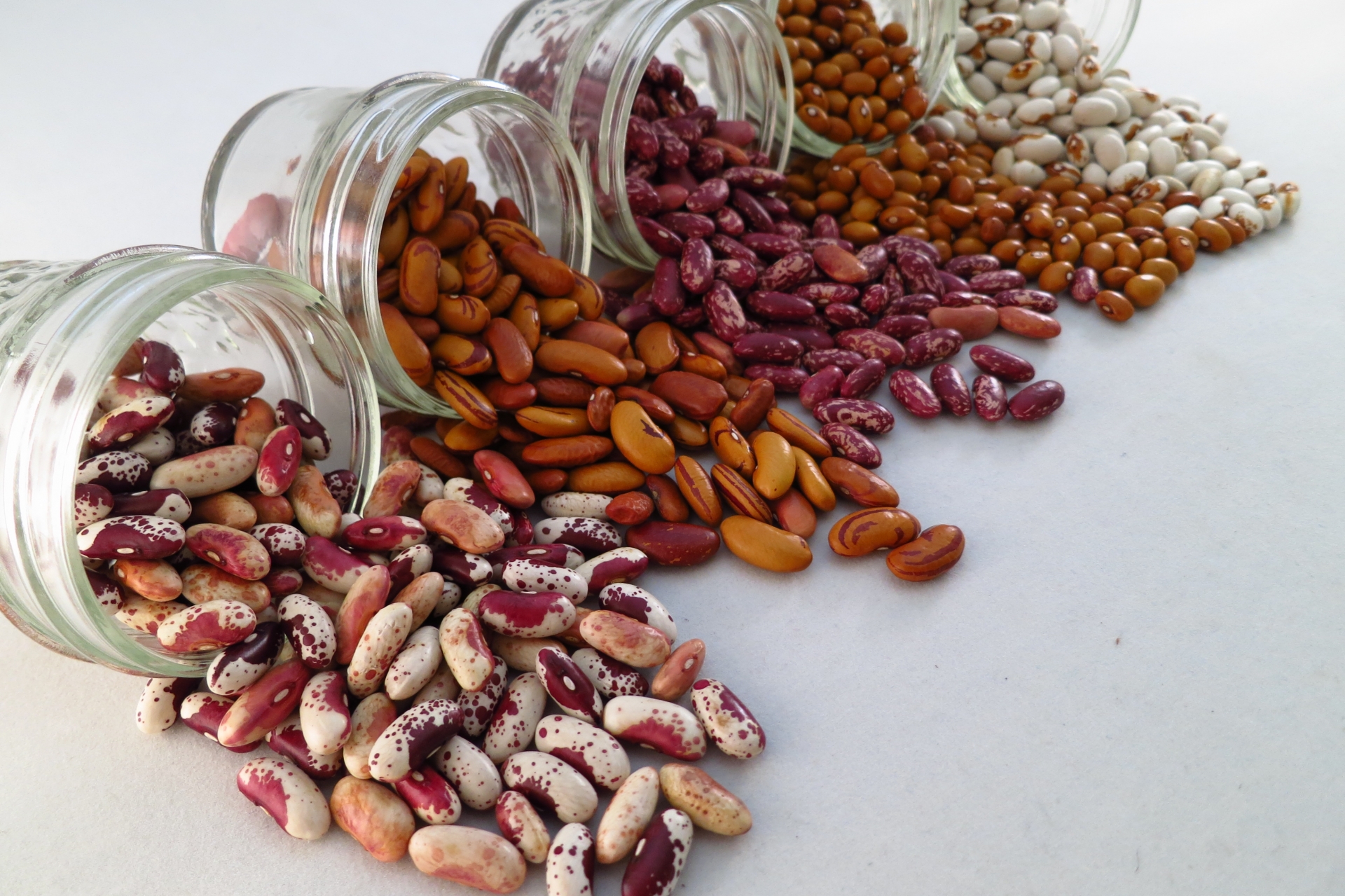 Four jars with different varieties of dried beans spilling out of them onto a white surface.