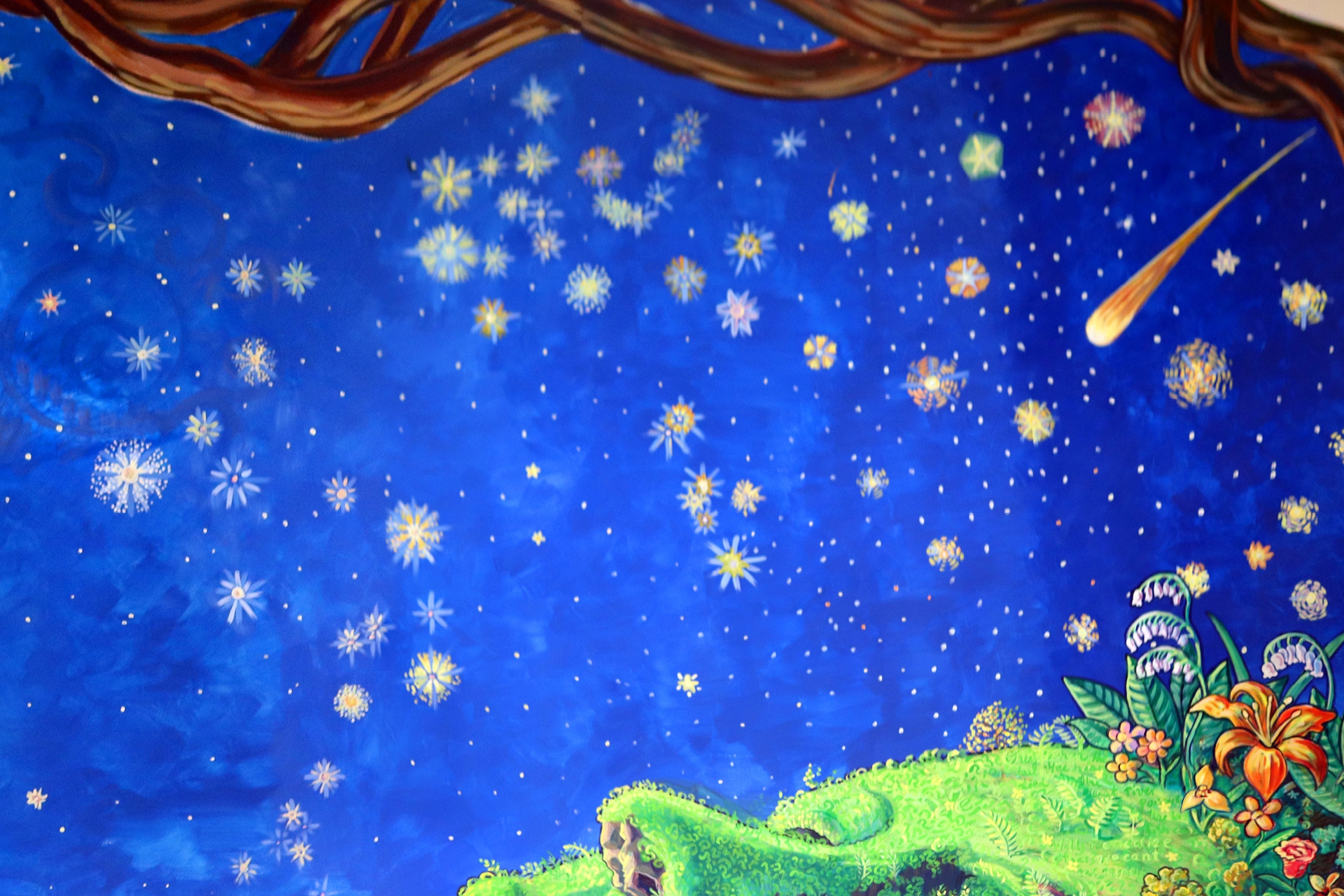 Stars painted on a rich blue background. In front of the background at the top of the picture there is a brown, gnarled tree branch. In front of the stars at the bottom of the picture there is a profile of a face looking up. The face is a hill, with grass for skin and flowers for hair. The face's eyes are closed.