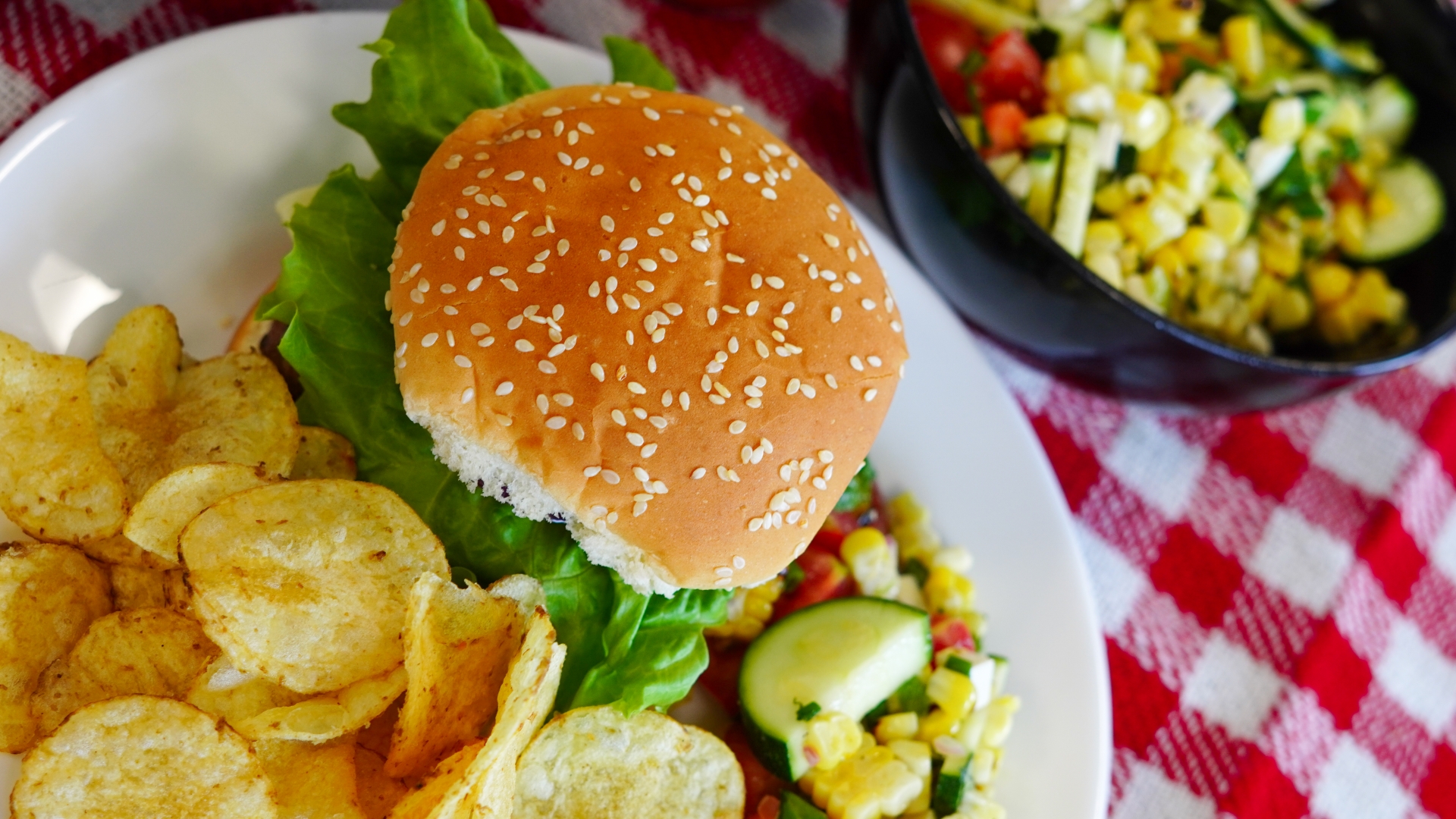 A top down view of a round white plate on a red and white checked tablecloth. There is a burger with a seeded bun and green lettuce sticking out the sides on the plate, along with a pile of yellow potato chips and a pile of corn and zucchini salad with tomato and herbs.