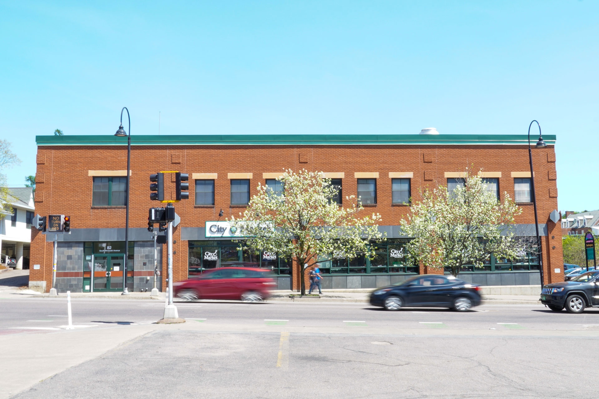 City Market's Downtown store viewed from street level, across South Winooski avenue. The building is rust red brick with green siding along the side of the roof. In front of the building, two trees are covered in white blossoms. A red hatchback and black sedan are on the street in front of the building. They are blurry due to movement.