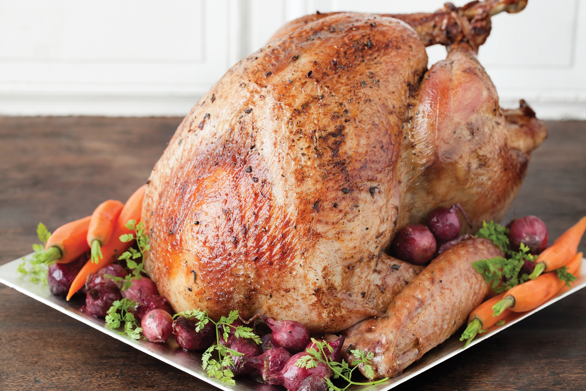 A roasted turkey on a platter with cooked carrots, small onions, and herb garnish. The platter rests on a dark wood counter with a white wall in the background.