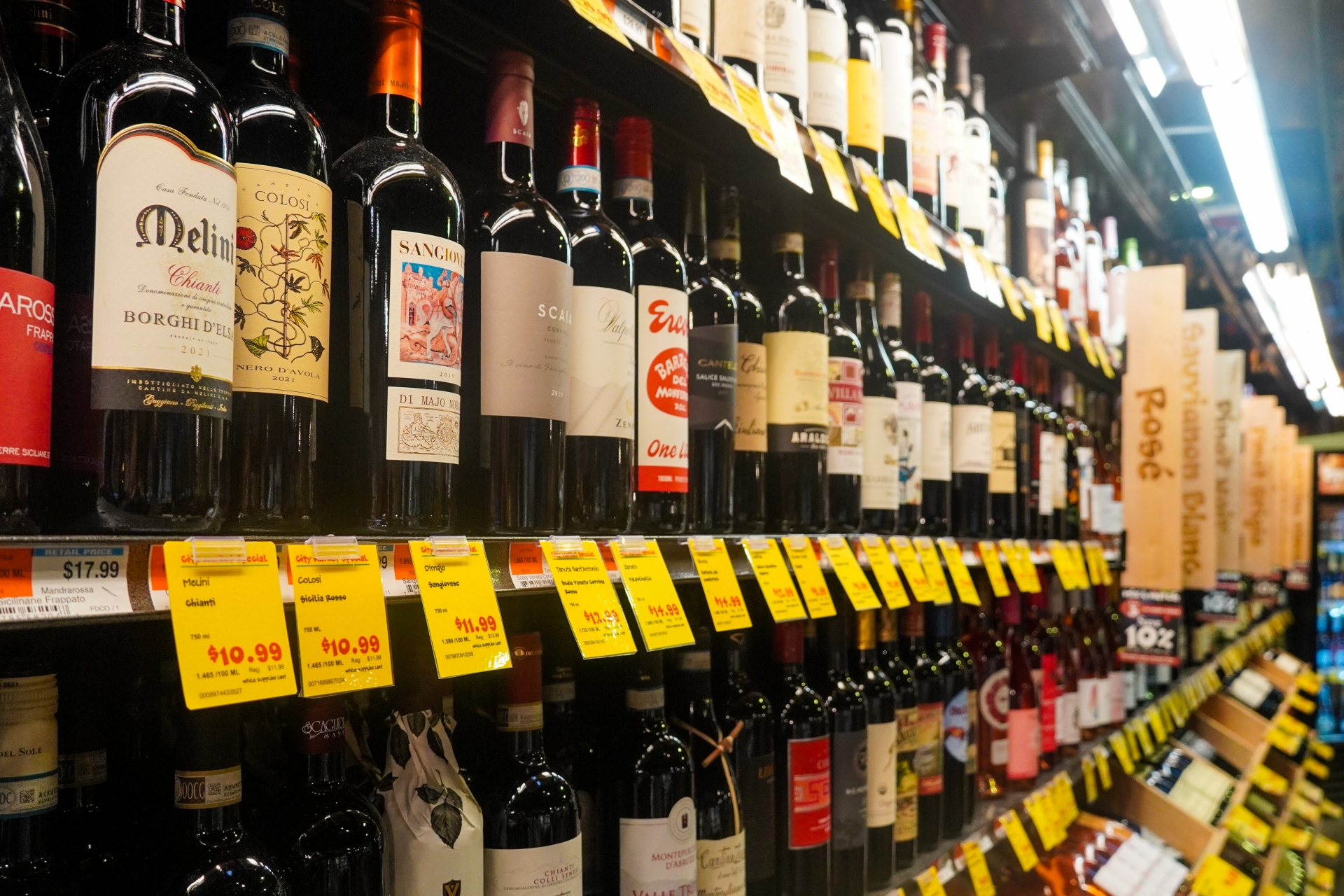 Bottles of wine with colorful labels displayed on several shelves in a store. Price tags run along the front of each shelf and vertical slats of wood with labels sub divide categories of wine.