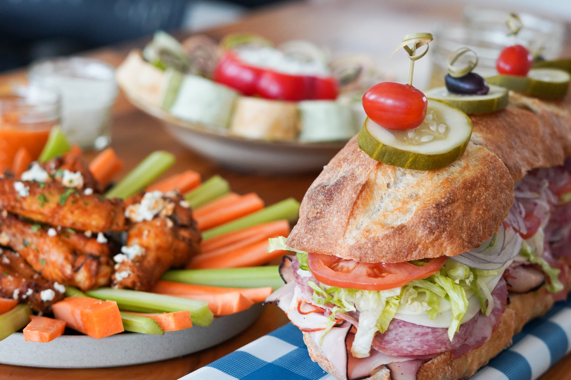 Several party foods are arrayed on a wooden table. In the foreground, a large sub sandwich on a crusty sub roll has sliced deli meats, shredded lettuce and a tomato slice peeking out the end. To the left, buffalo wings with blue cheese crumble sit at the center of a platter, surrounded by carrot and celery sticks. In the background, an additional platter holds sliced sandwich pinwheels and veggies.