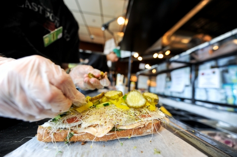 A close up of a person's hand in a latex glove placing green pickle rounds on an open sandwich that features sprouts, cucumber, tomato, sliced cheese and sliced turkey. The sandwich is on a light countertop.