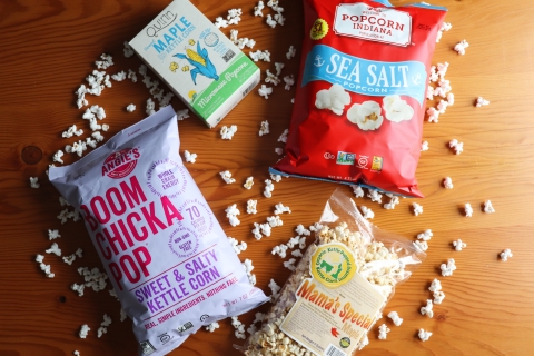 Popped popcorn scattered on a wooden table with three unopened popcorn packages. The first is a white box with a drawing of an ear of corn on it. The second is a lavender bag labeled "Boom Chicka Pop." The third is a red bag labeled "Popcorn Indiana." The last is a clear bag with popped popcorn visible inside, labeled "Mama's Special."