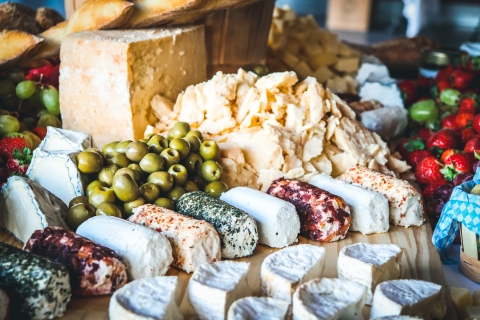 A cheese spread with a section of a large creamy-yellow hard cheese wheel. In front of the cheese wheel are a pile of crumbled hard cheese and a pile of olives. In front of the olive pile are nine rolls of chevre, crusted with a variety of colorful herbs and dried fruit. In front of the chevre are seven half-moons of soft cheese with white rinds. Behind the display are bright green grapes and bright red strawberries.