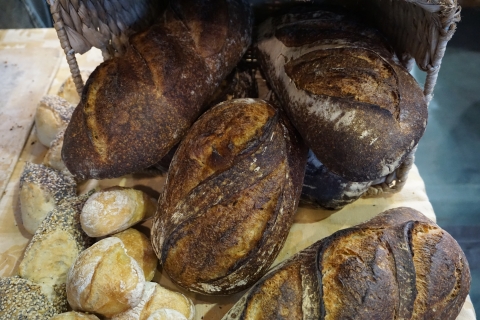 Four large, dark loaves of bread on a light wooden table, with several golden brown rolls to the left-hand side. The left-most rolls are seed-crusted, and the rest are dusted with flour.