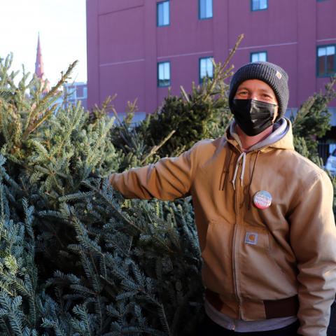 A person in a brown coat and a gray winter hat holds a pine tree. There is a pin on his coat that says "Member Worker."