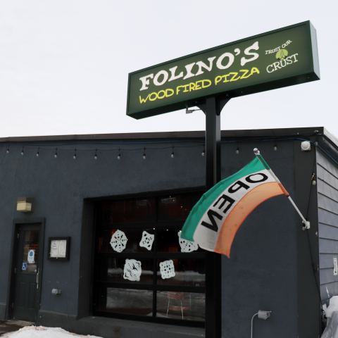 The exterior of a one-story dark gray building. There is a glass door to the far left and a large window on the right with paper cut-out snowflakes scattered over it. The sign above the building is green and has "Folino's Wood Fired Pizza" printed on it in white and yellow text." There is a green, white and orange "open" flag flying from the corner of the building.