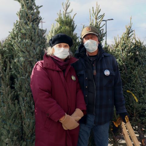 Two people stand in front of a row of cut pine trees. The person on the left is wearing a long maroon puffy coat, brown knit gloves and a hunter green felt cap. The person on the right is wearing an olive green canvas cap with a brown patch on the front, a blue and black plaid wool coat, black gloves, and jeans.