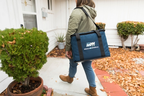 A person walks on a white path lined with bricks. They are wearing a gray coat, jeans and brown boots and they are carrying a blue bag with a white logo that reads "Meals on Wheels." The person is passing a green potted plant on their left and approaching the door to a white house.
