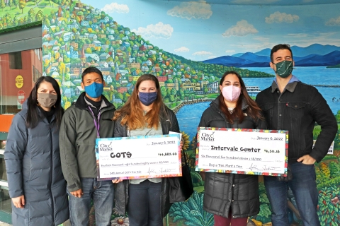3 City Market Staff Members are posing with a COTS staff person and an Intervale Center Staff person. The COTS staff person and the Intervale Staff person are holding oversized checks. They are standing in front of a mural of a lake.
