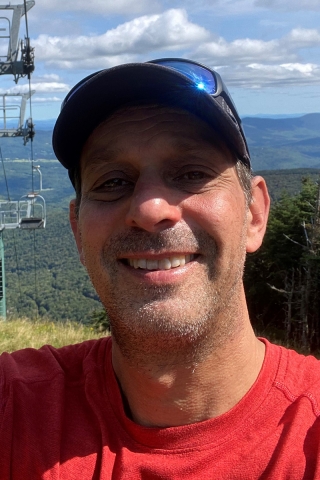 A portrait of John Davidowitz in a red t-shirt with a baseball hat and sunglasses on top of his head, standing outside with a ski lift and mountains behind him