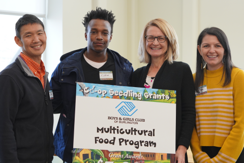 Four people in a row pictured from the waist up. The two people in the middle are holding a sign with the Boys & Girls Club of Burlington logo and the words "Multicultural Food Program" printed on it in black lettering. The top of the sign reads "Co-op Seedling Grants."