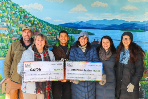 Six people in winter coats stand in a row in front of a painted mural. The mural features a mountain on the left and a lake on the right. The second and fourth people from the left each hold an oversized check. The leftmost check is made out to COTS. The rightmost check is made out to the Intervale Center.