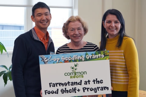 Three people in a row pictured from the waist up. The person in the middle is holding a sign with the Common Roots logo and the words "Farmstand at the Food Shelf+ Program" printed on it in black lettering. The top of the sign reads "Co-op Seedling Grants."