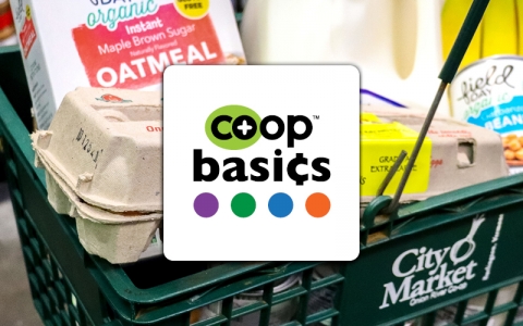 A square logo on a white background that reads "Co-op basics" in black lettering. Below the lettering there are four small circles in a horizontal row. The first is purple, the second is green, the third is blue and the fourth is orange. The logo is in front of a photo of a green City Market shopping cart filled with groceries - a box of instant oatmeal, a carton of eggs and a gallon of milk.