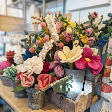 Felted flowers arranged in tin metal buckets on a wooden display table. The flowers are a variety of shapes and colors - some are tall with small white flowers, others have wide pink petals or yellow small petals.